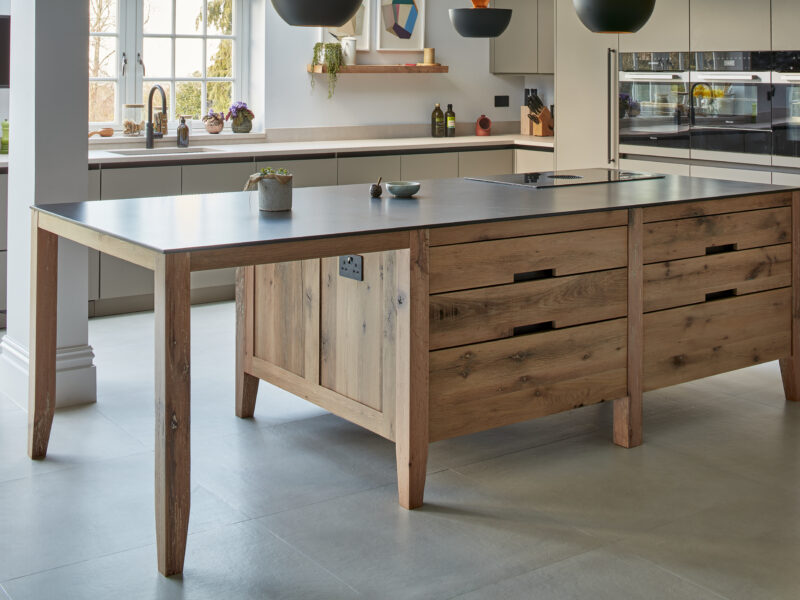 Dark-stained oak island with pale grey worktop, sage cabinetry and black pendant lights by a garden window