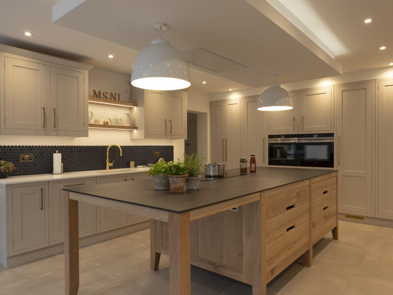 Light oak island with grey worktop in a neutral kitchen of cream units and dark pendants
